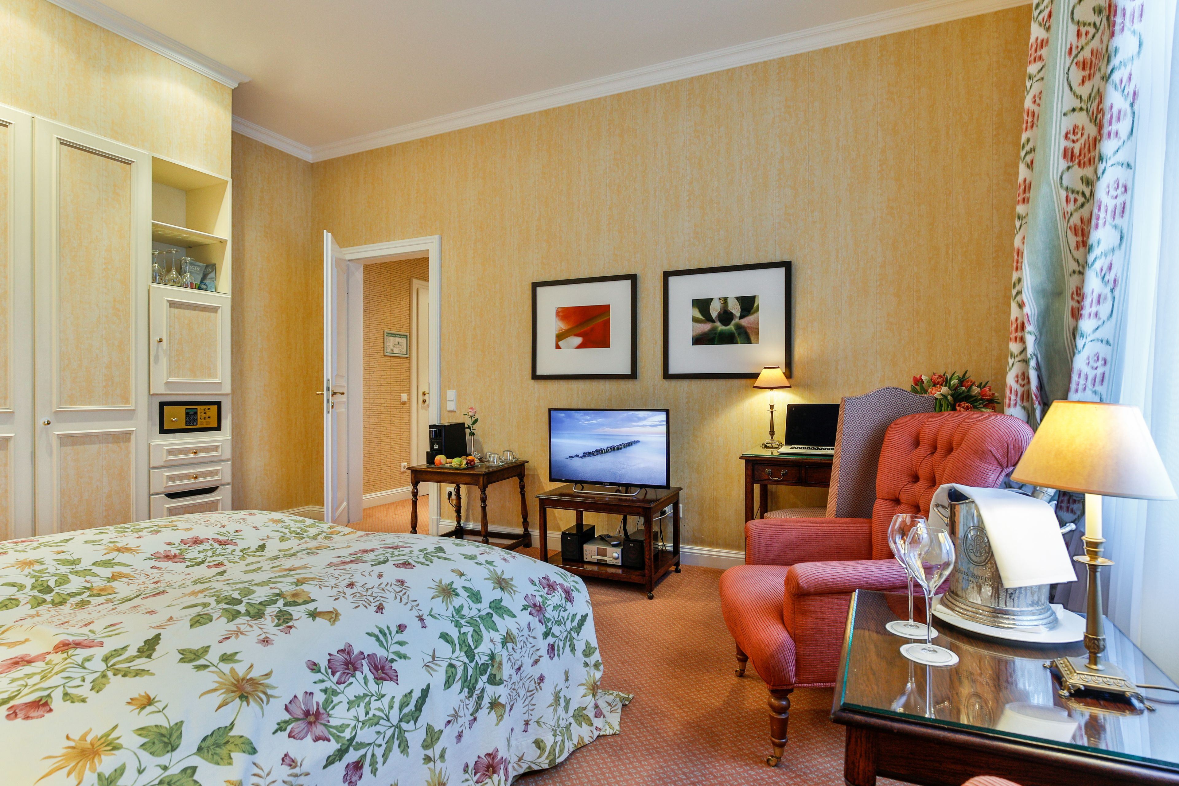 Una habitación de hotel con una cama, televisión, una silla y una mesa con lámpara y copas de vino.