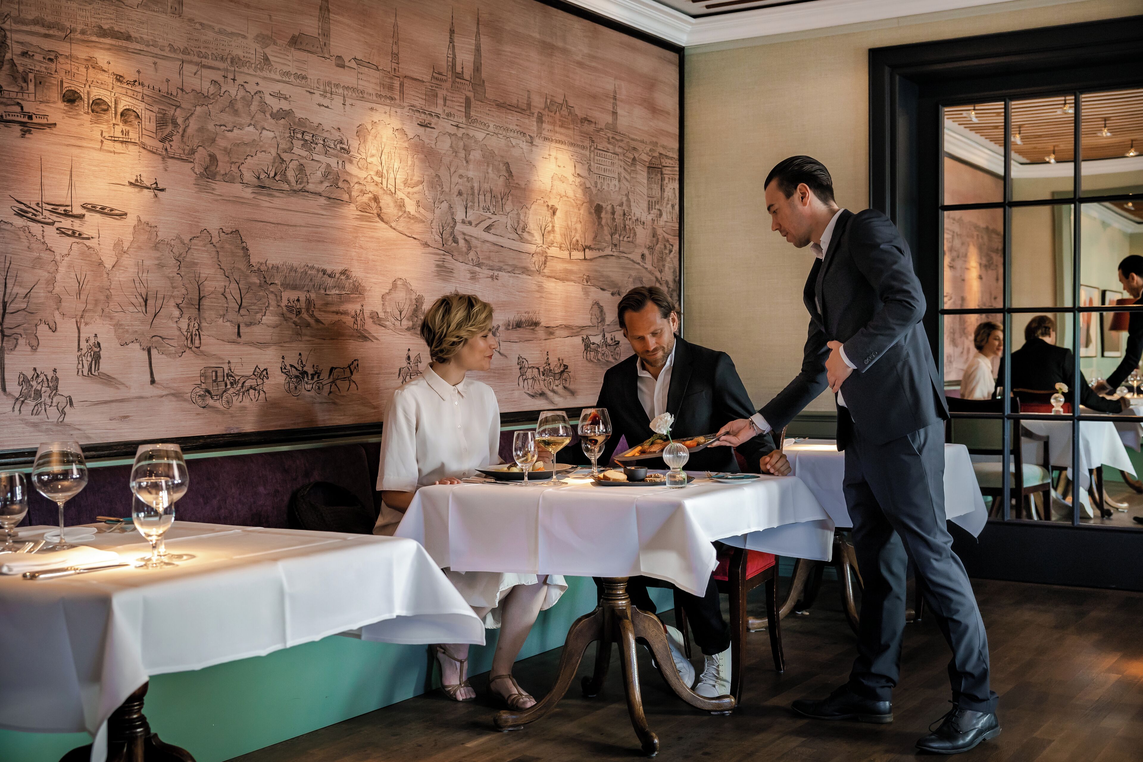A restaurant with a mural of a cityscape on the wall, tables set with wine glasses and plates, and a waiter attending to guests.