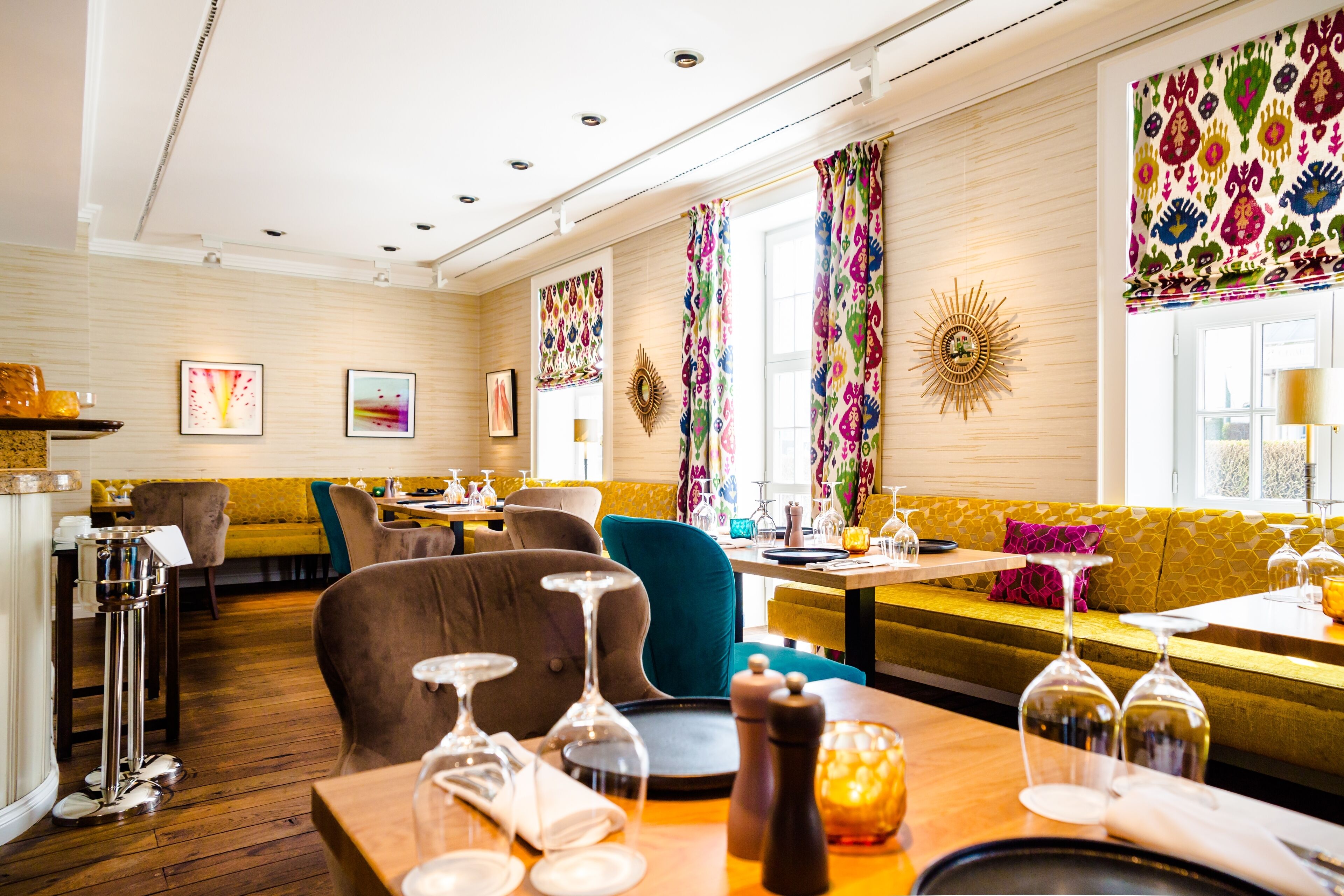 A restaurant with wooden floors, multiple seating areas including a long table and a sofa, and colorful patterned curtains.