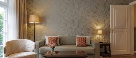 A living room with a sofa, armchair, and coffee table, featuring patterned wallpaper and a door.
