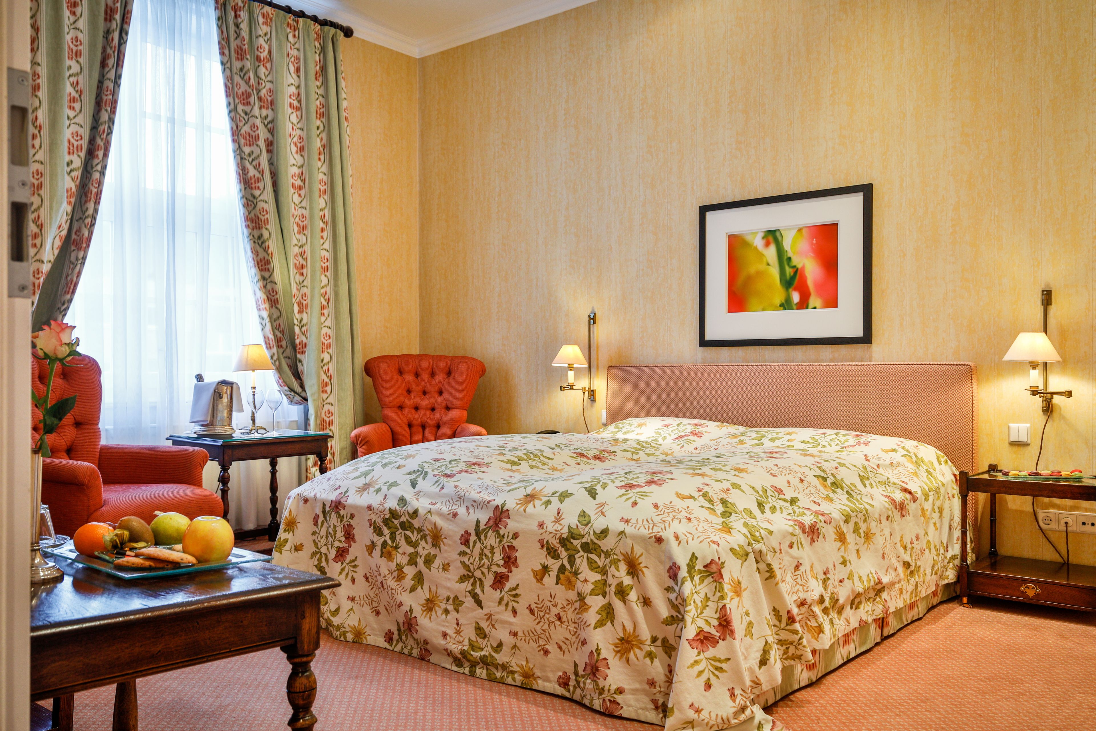 A hotel room with a floral bedspread, a red armchair, a table with fruit and a vase, and a framed picture on the wall.