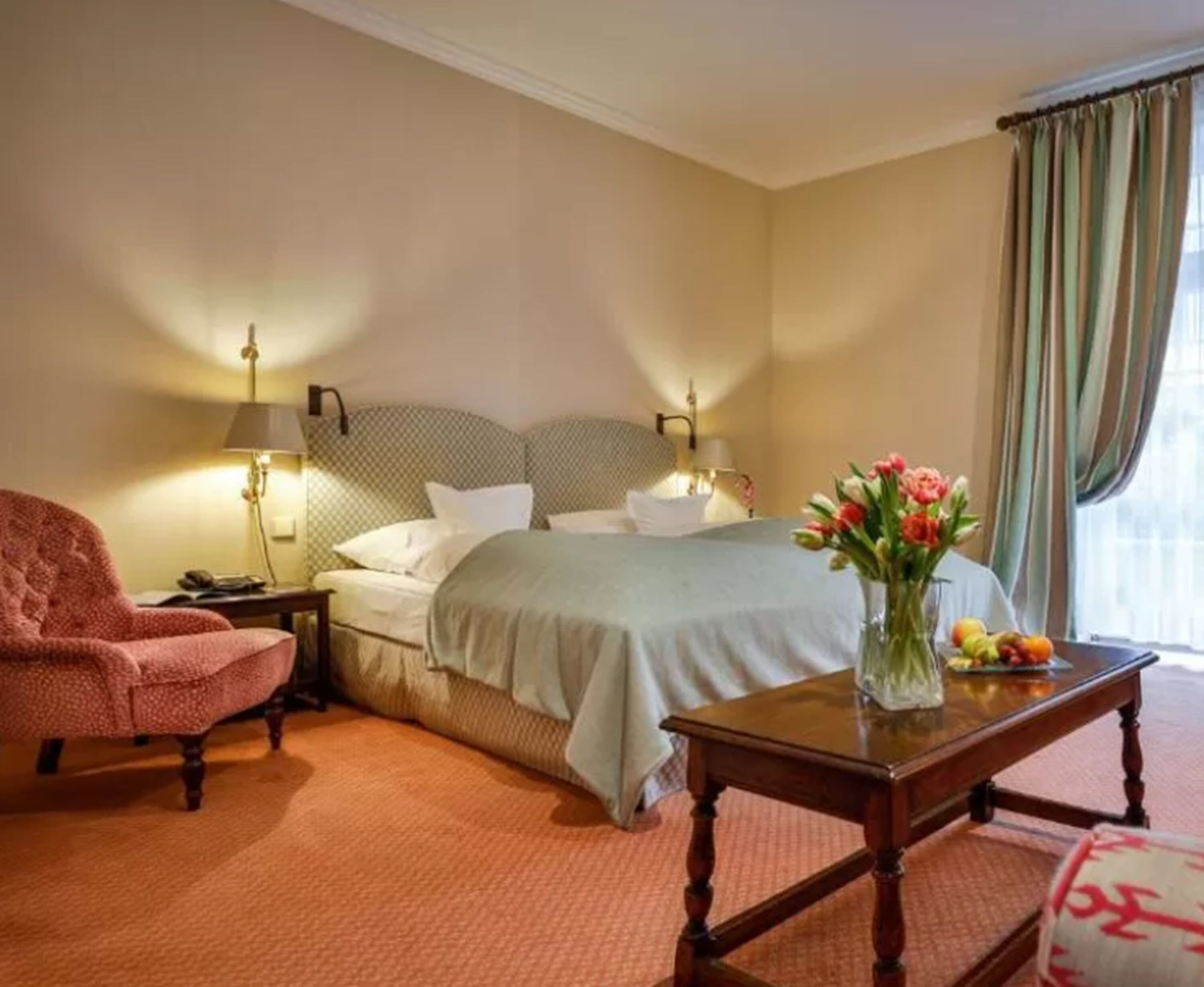 A hotel room with a bed, a chair, a nightstand, a lamp, a table with a vase of flowers, and a window with curtains.