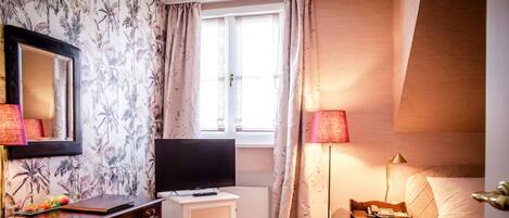 A hotel room with a bed, a TV, a desk with drawers, a lamp, and a window with curtains.