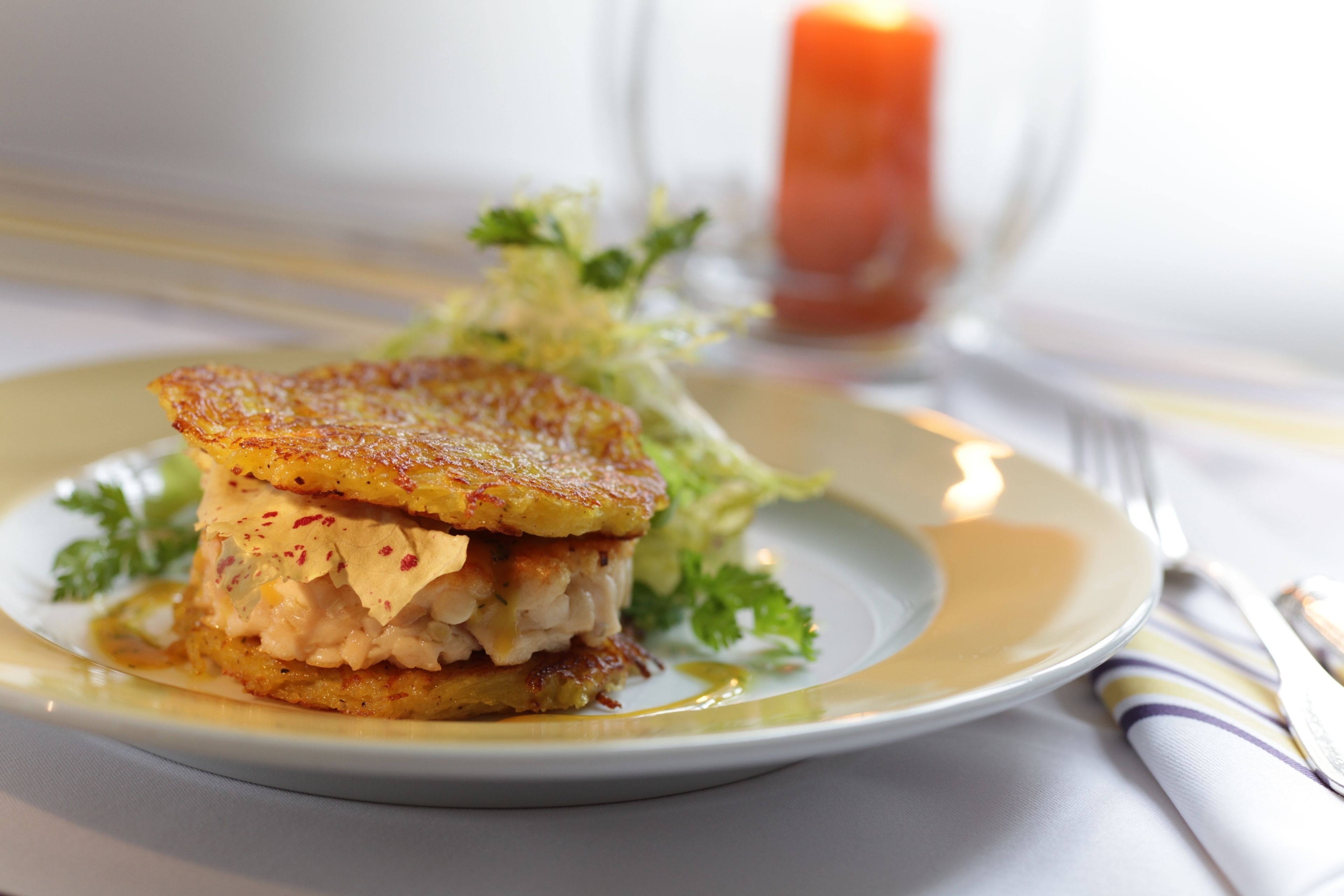 A plate with a food item, possibly a sandwich or a layered dish, with a side of greens and a small candle in the background.