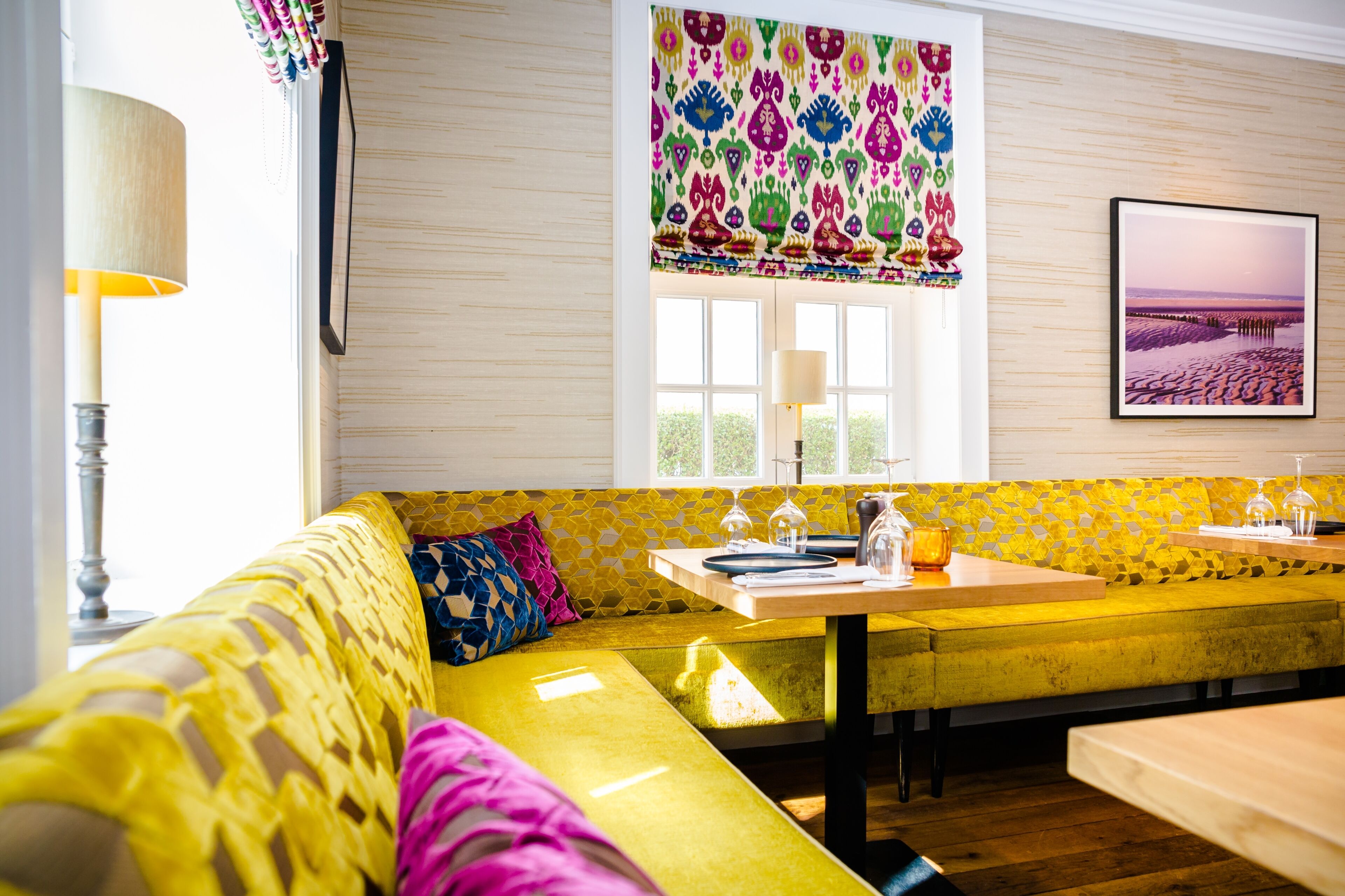 A bright yellow sofa with a bench, a table with a lamp, and a window with a colorful valance.