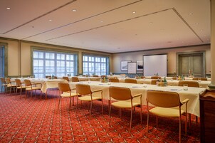 Un salón de conferencias con varias mesas y sillas, ventanas amplias y una pantalla de proyección.
