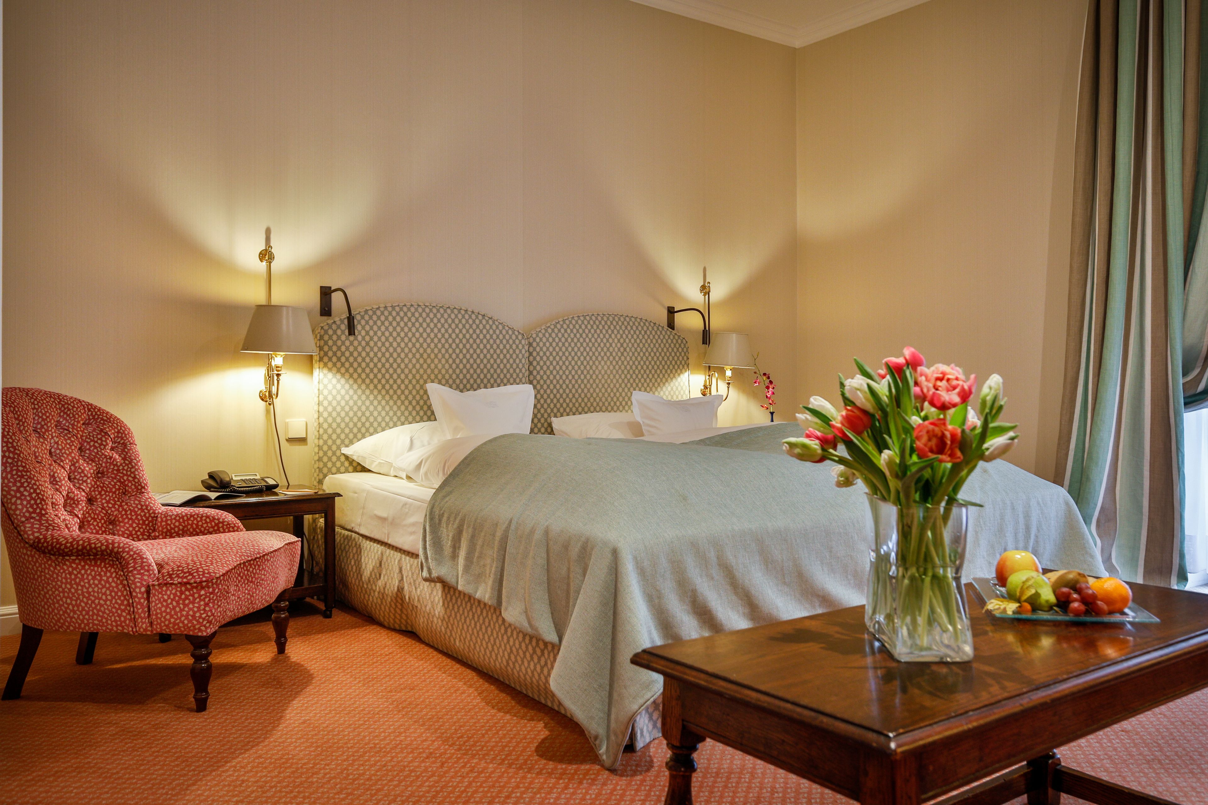 Ein Hotelzimmer mit Bett, Stuhl, Nachttisch, Blumen in einer Vase und Obstkorb.