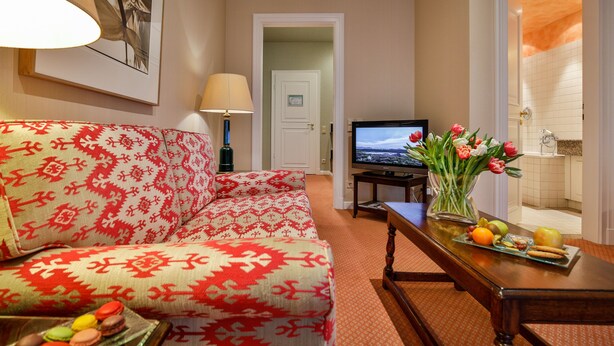 A living room with a patterned sofa, a television, a table with fruit and flowers, and a door leading to a bathroom. - Hotel Stadt Hamburg (Sylt)