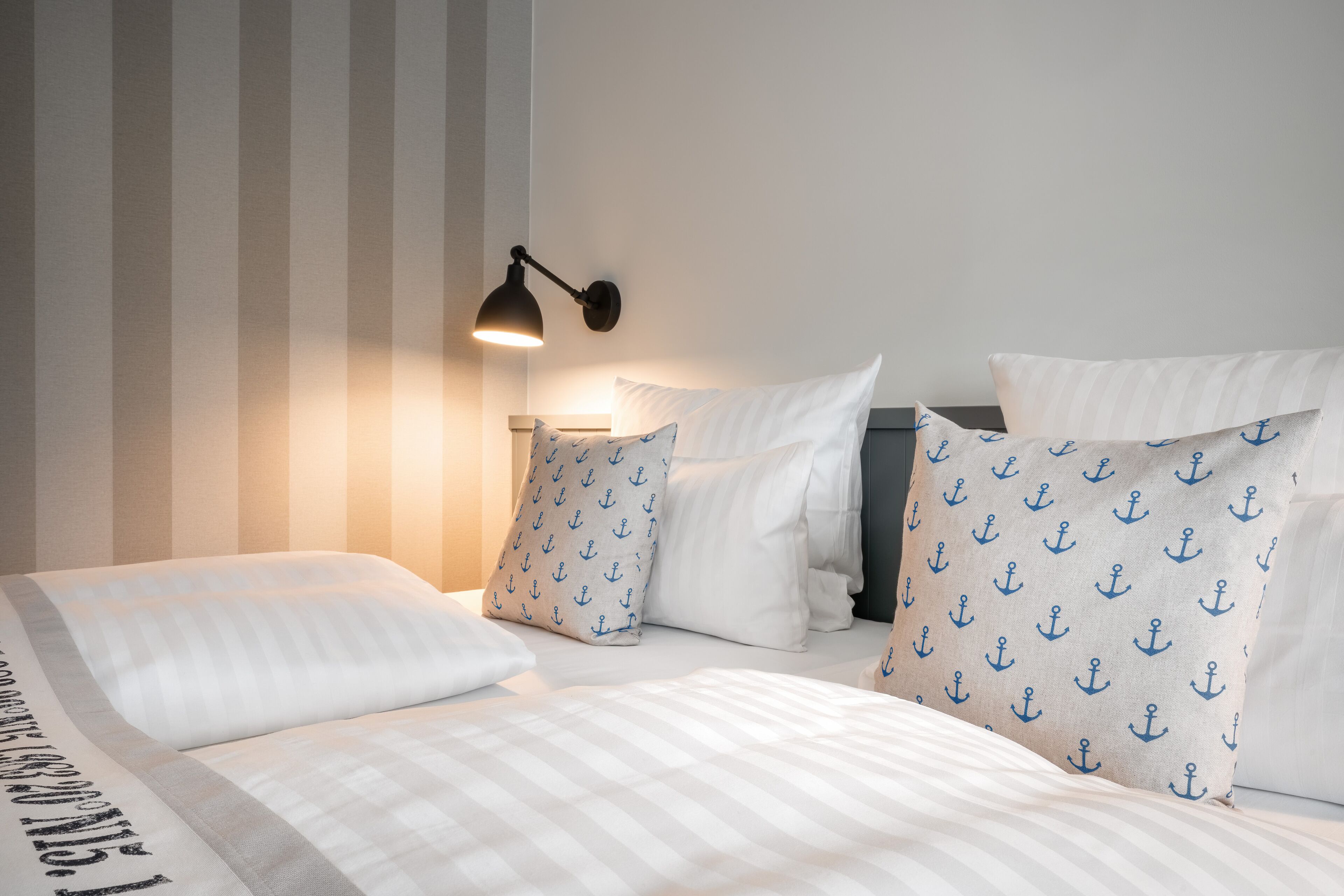 Una cama bien hecha con ropa de cama a rayas, una lámpara de pared y almohadas con estampados de anclas.