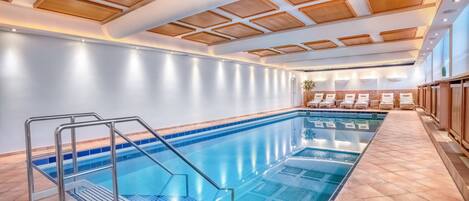 En swimmingpool med en metallstege och solstolar, omgiven av ett kaklat golv och träpaneler i taket.