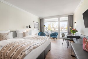 Quarto de hotel com uma cama grande, uma escrivaninha, duas cadeiras, uma mesa pequena e uma janela com vista para o exterior.