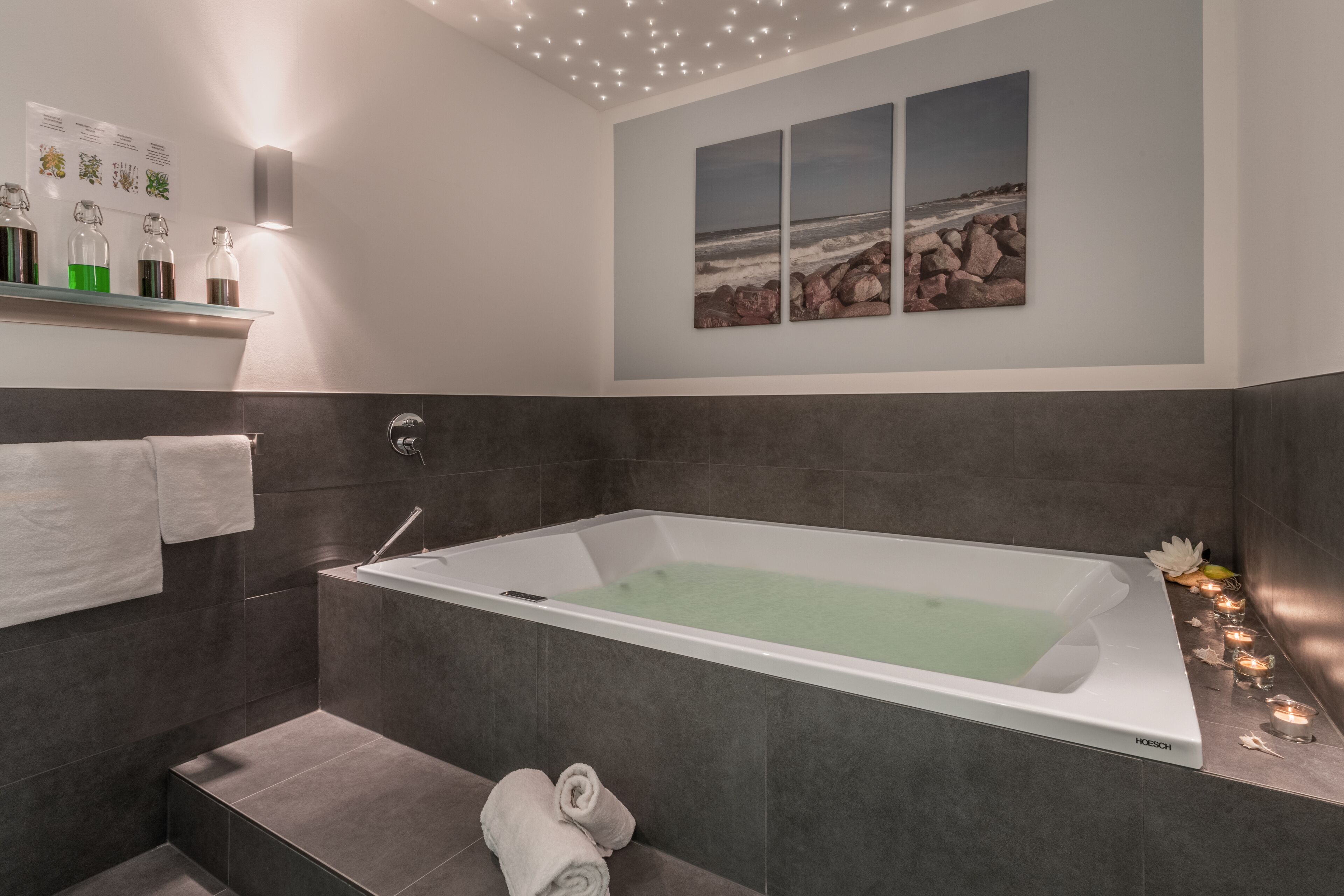 Un bagno moderno con una vasca da bagno spaziosa, piastrelle scure, un porta asciugamani e uno scaffale a parete con flaconi.