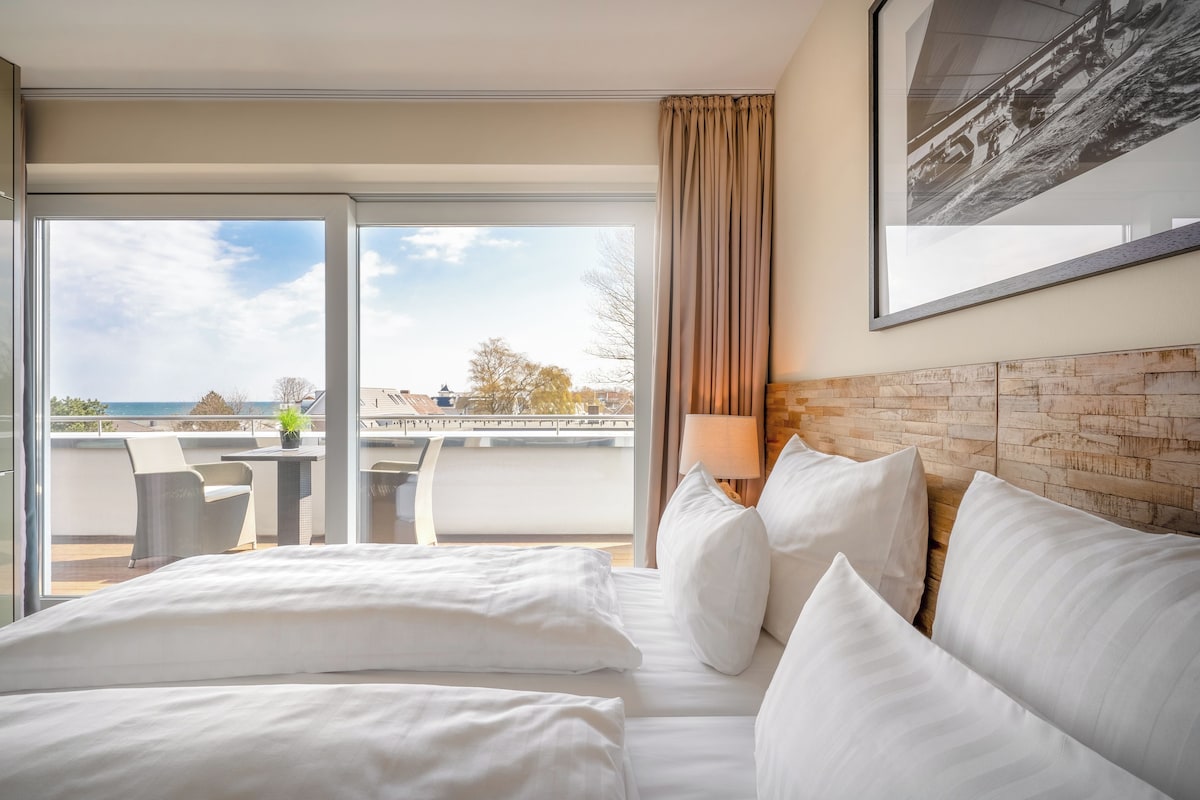 Een hotelkamer met een groot bed, een balkon met een tafel en stoelen, en uitzicht over de stad.