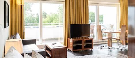Habitación de hotel con cama, televisor sobre un soporte de madera, escritorio con silla y balcón con cortinas amarillas.