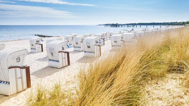 Reihe nummerierter Strandkörbe entlang eines Sandstrandes, im Hintergrund ein Steg.