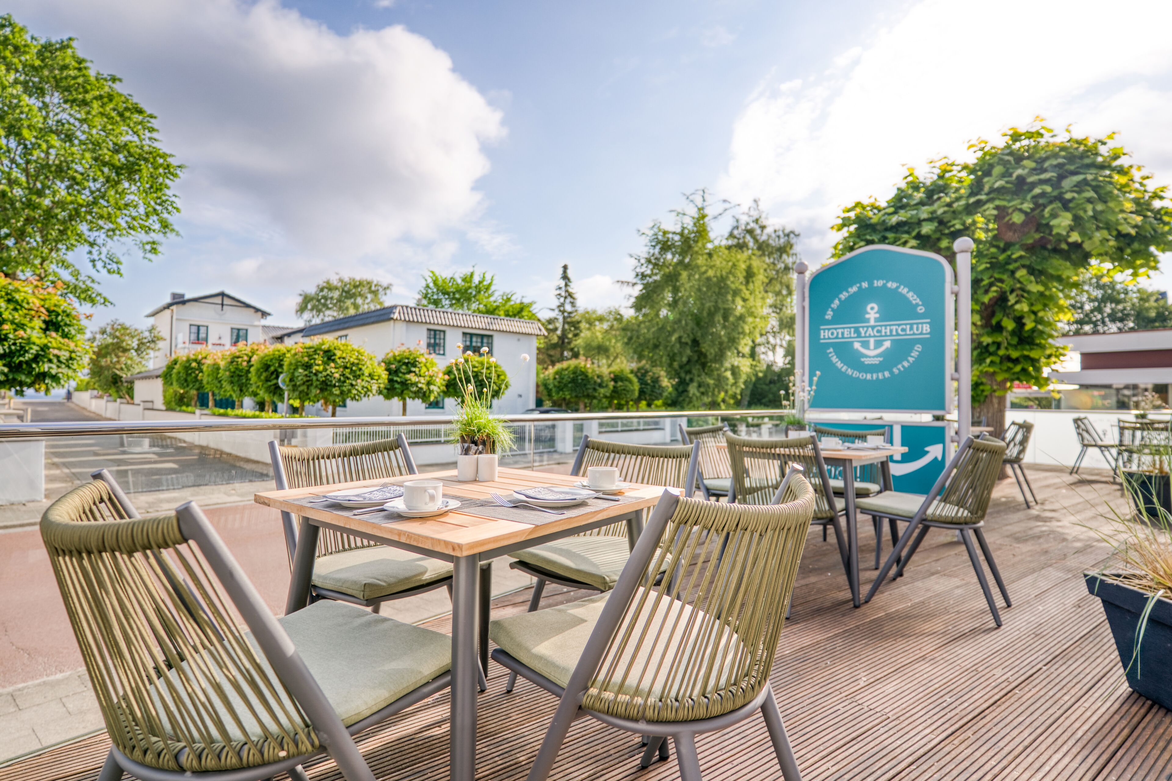 Un espace extérieur aménagé pour les repas, avec des chaises et des tables en osier, une enseigne indiquant l’hôtel Yacht Club, et une vue sur les bâtiments et les arbres.