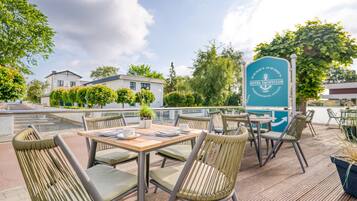 Een terras met rieten stoelen en tafels, een bord met Hotel Yacht Club erop, en uitzicht op gebouwen en bomen.