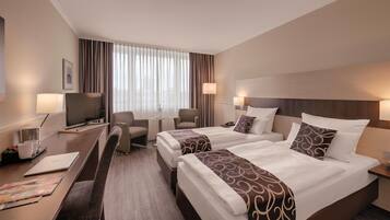 A hotel room with two beds, a desk with a laptop, a chair, a TV, and a window with curtains.