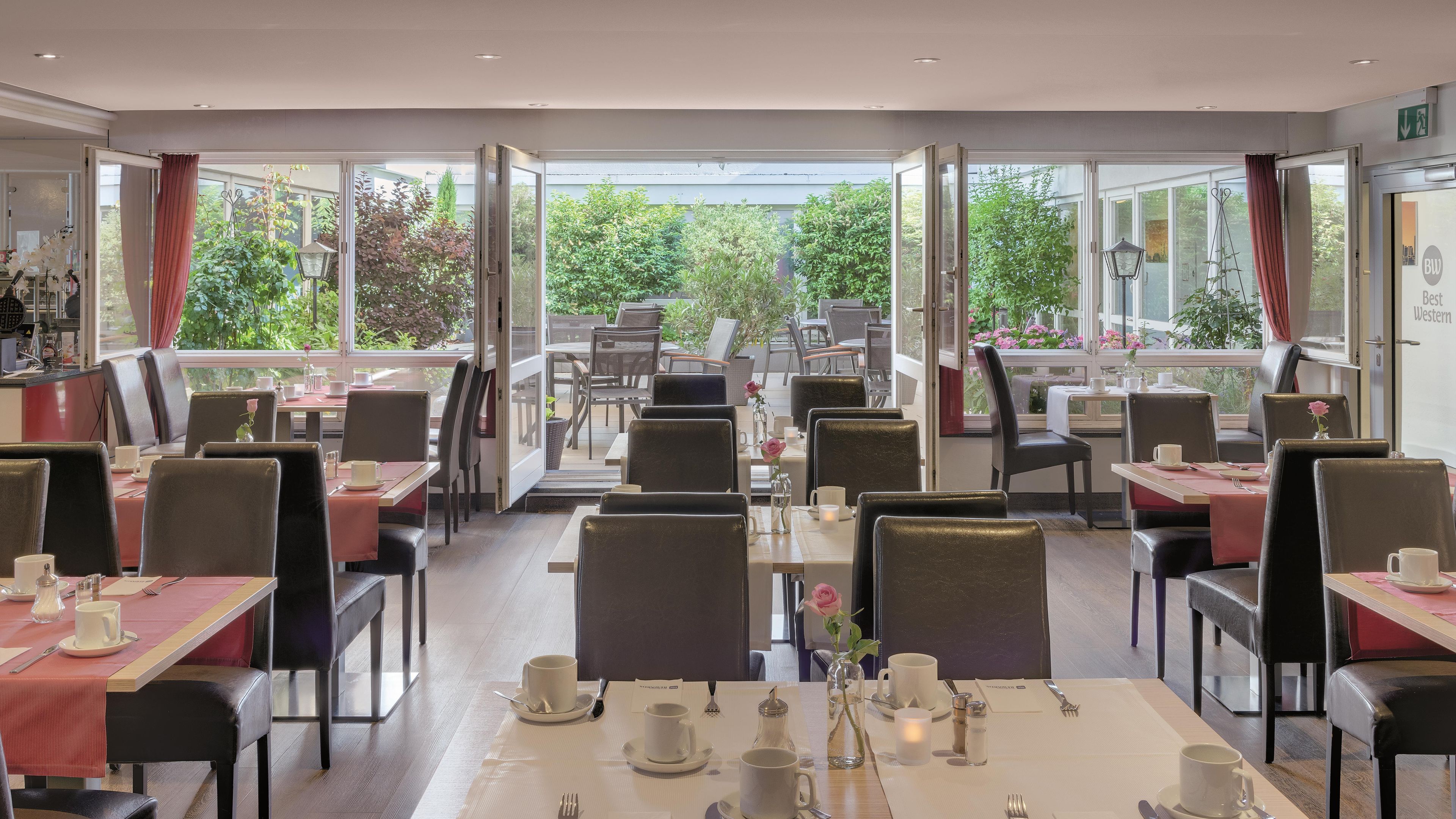 A restaurant with tables set for dining, a view of greenery outside, and a sign indicating 'Best Western'.