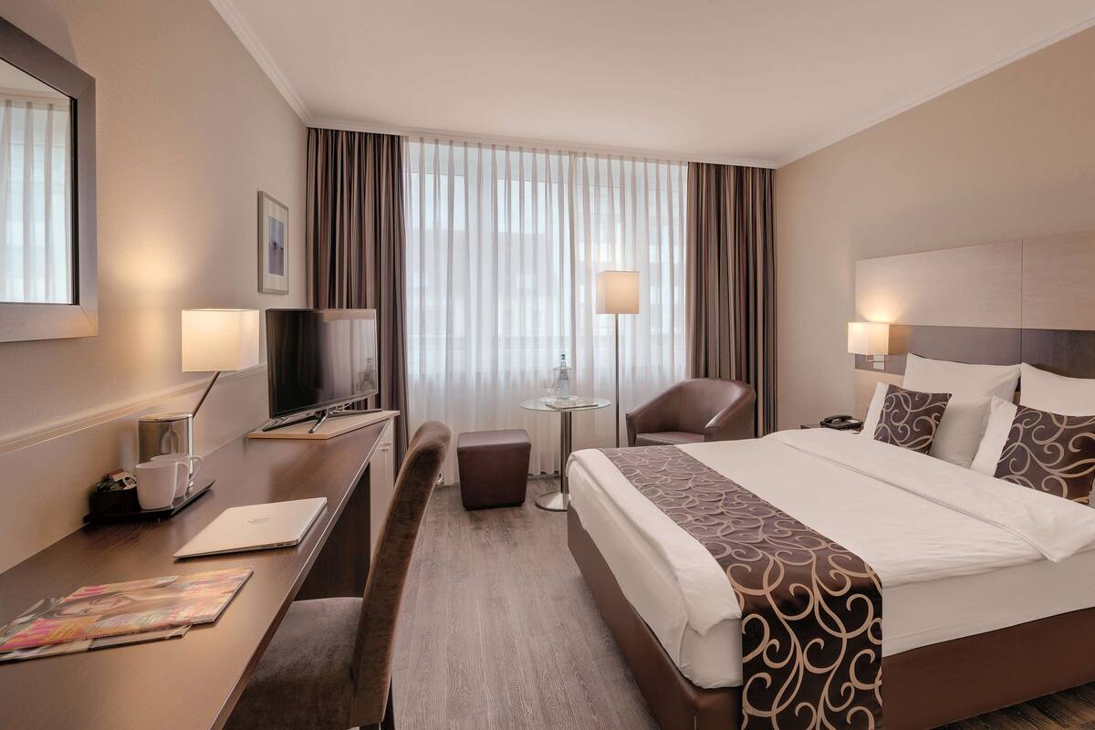 A hotel room with a large bed, a desk with a computer, a chair, a TV, and a window with curtains.