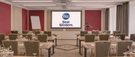 Un salón de conferencias con una pantalla de proyector mostrando la marca 'Best Western', varias mesas con sillas y una pizarra.