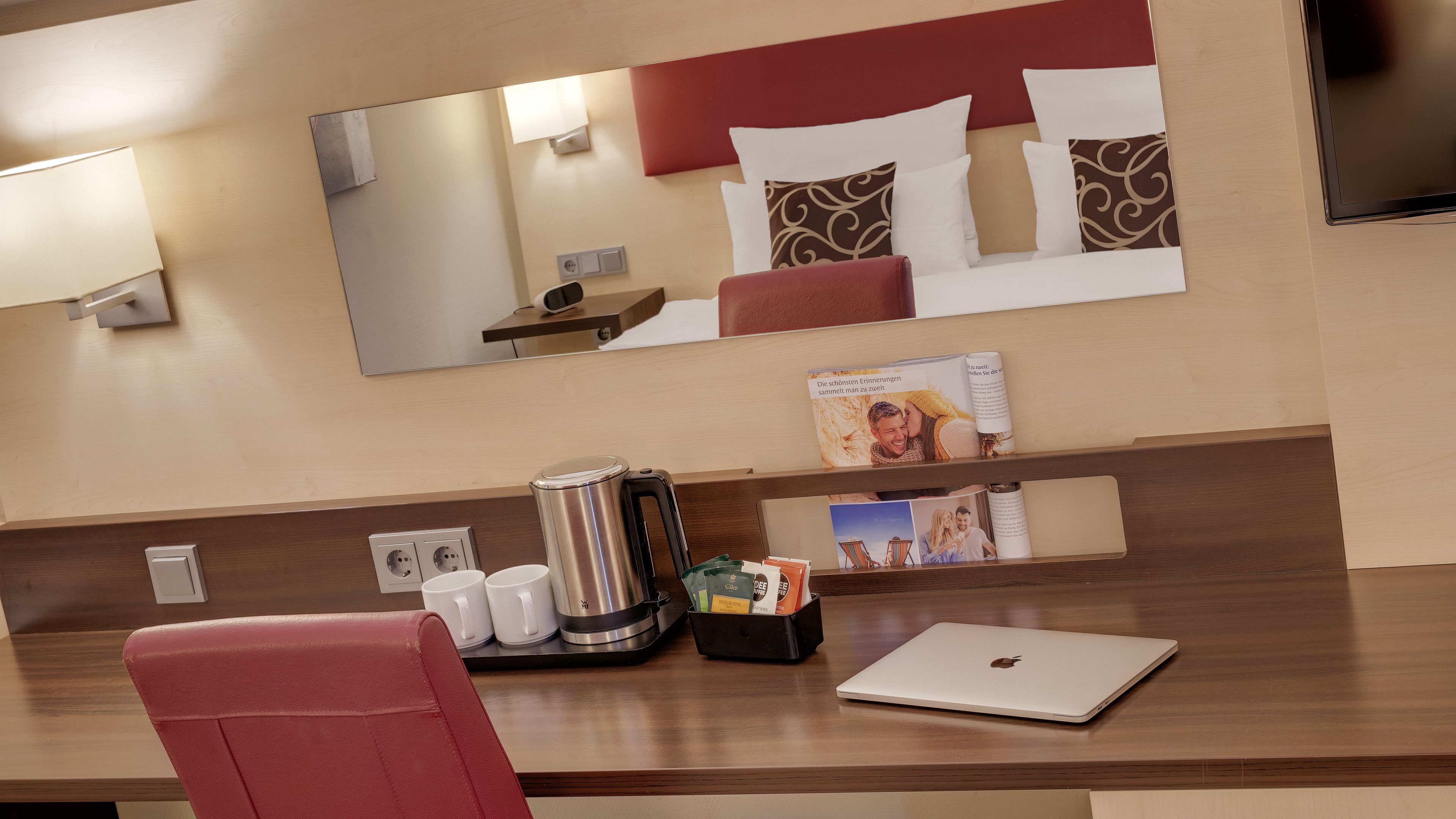 Een hotelkamer met een houten bureau, een rode stoel, een laptop, een koffiezetapparaat en een tijdschriftrek met boeken en tijdschriften.