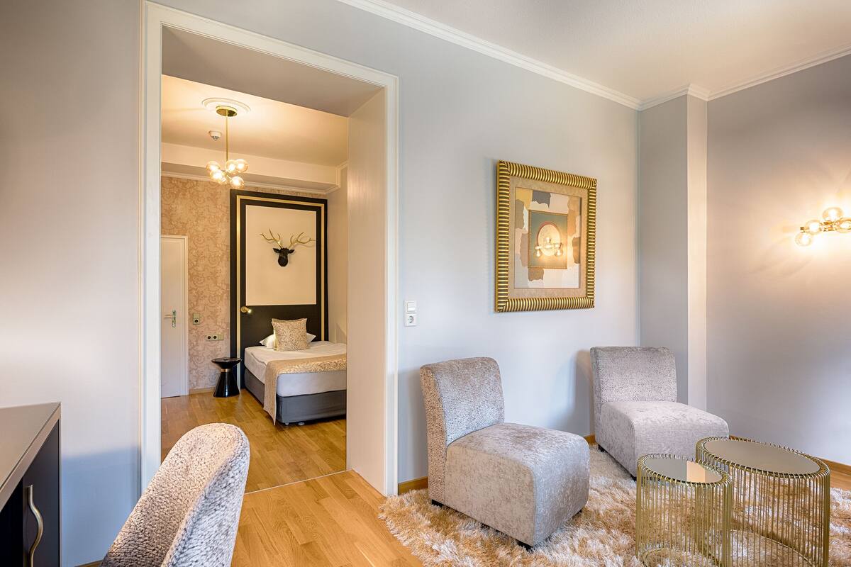 Una sala de estar moderna con un sofá, sillones y una mesa de centro. Hay un espejo en la pared y una puerta que da a otra habitación.
