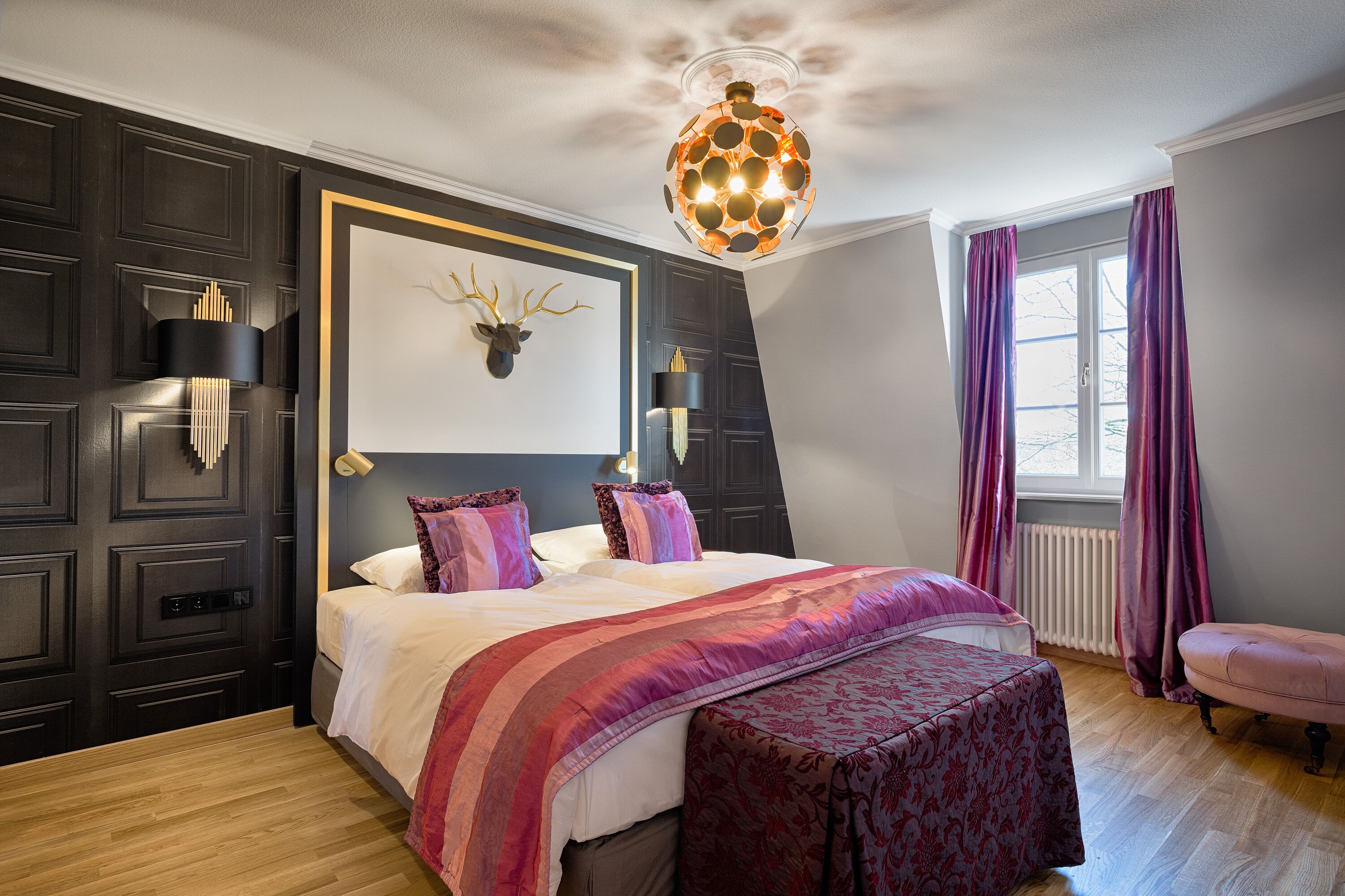 Un dormitorio con una cama grande, una lámpara decorativa en el techo, una cabeza de ciervo colgada en la pared y una ventana con cortinas.