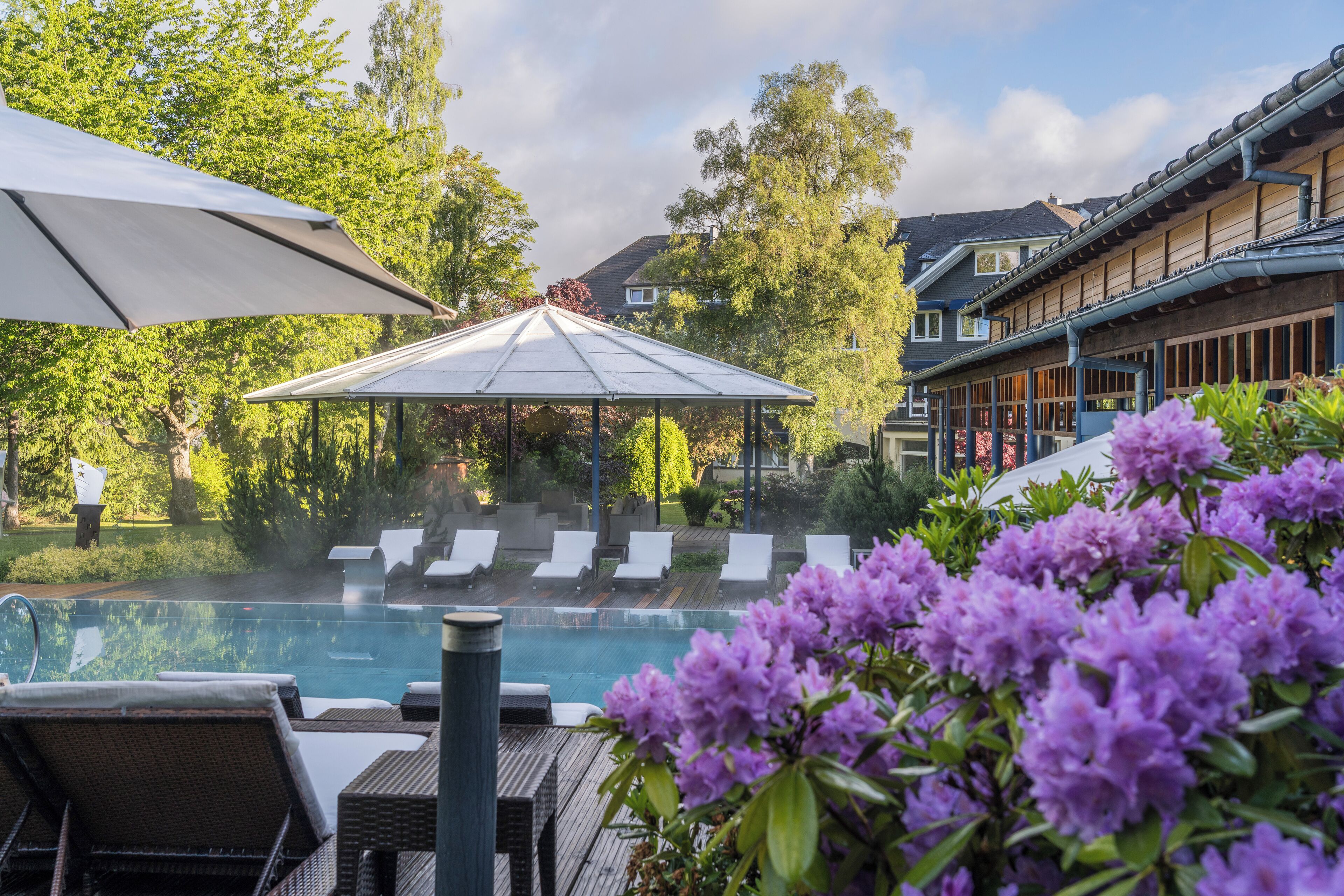 Un espace piscine avec des chaises longues, un kiosque et des fleurs violettes.