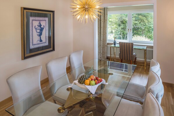 A dining area with a glass table, white chairs, a fruit bowl, a framed artwork, and a window with a view of trees. - Parkhotel Adler (Hinterzarten)