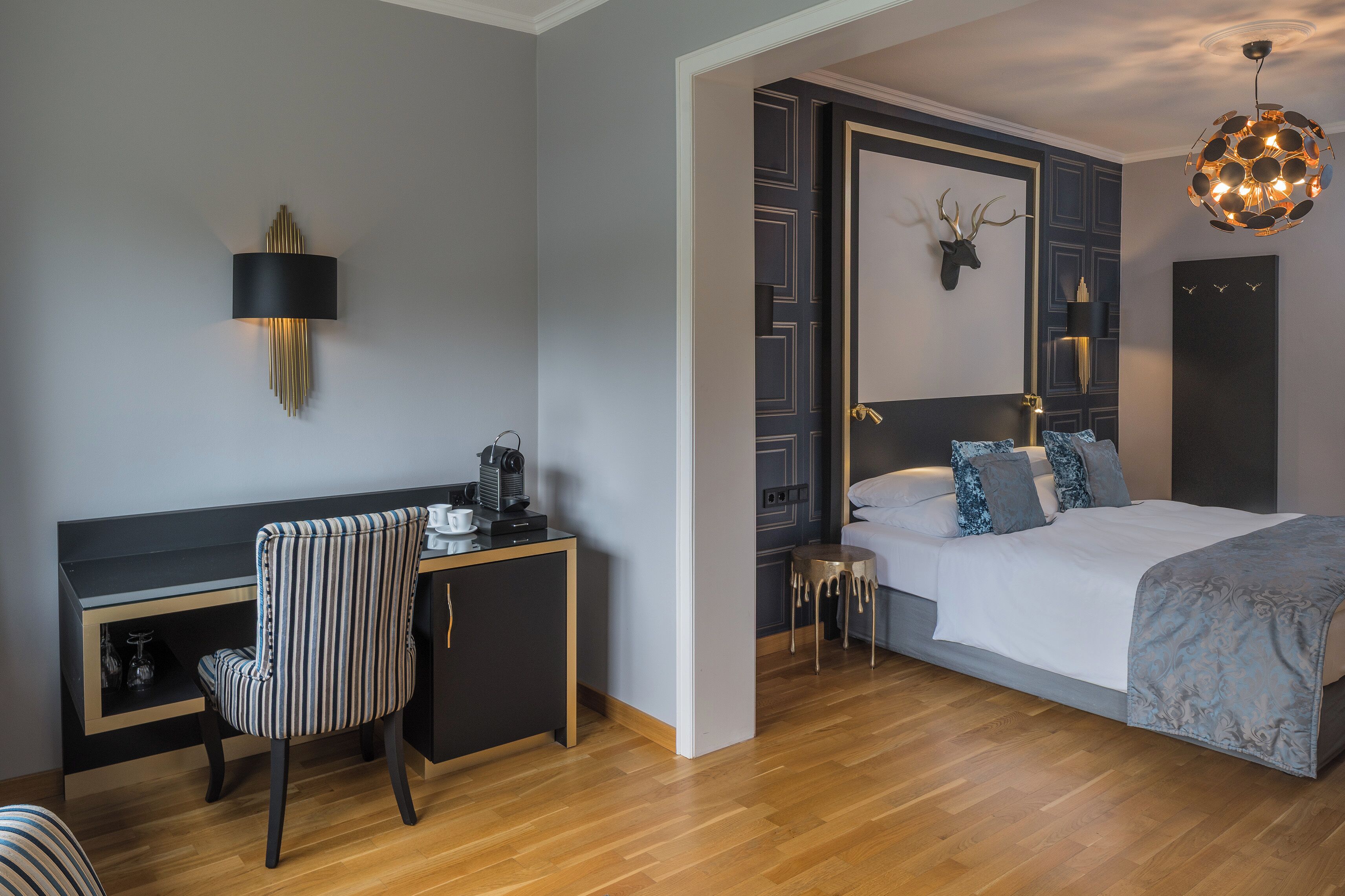 A modern hotel room with a wooden floor, a desk with a chair, a bed with a headboard, a wall-mounted lamp, and a unique ceiling light fixture.