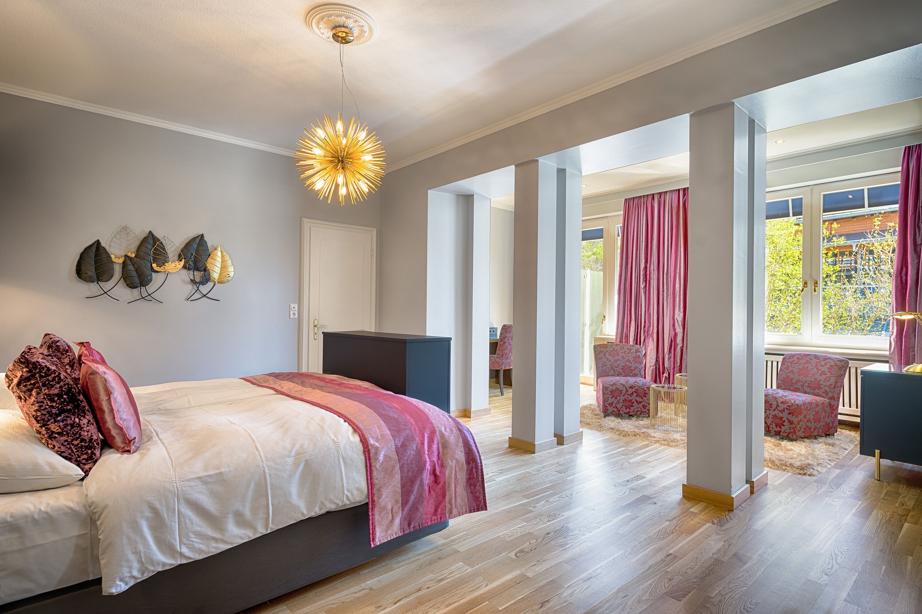 A modern bedroom with a large bed, a TV, and a unique ceiling light. There are two chairs and a window with curtains.