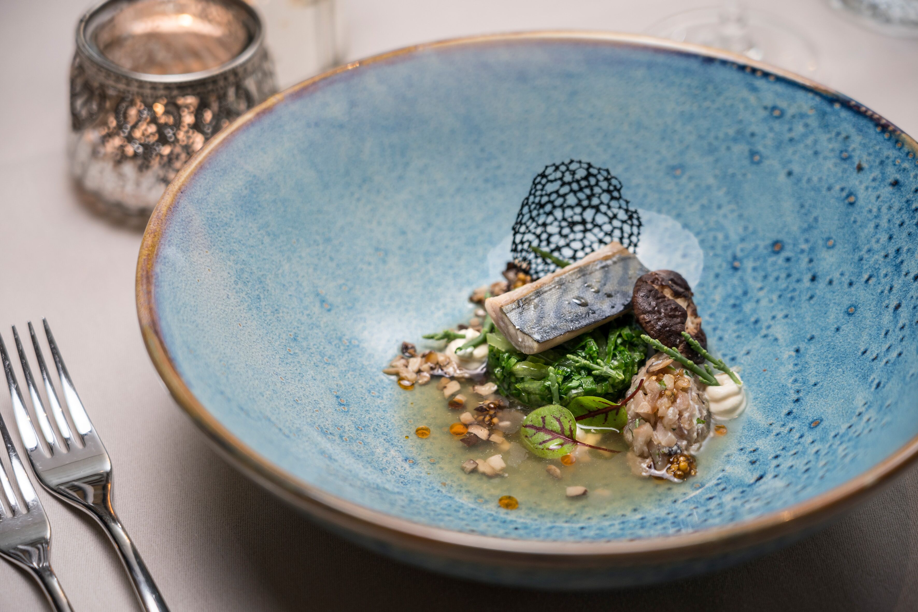 A blue ceramic plate with a fish, greens, and nuts on a table.