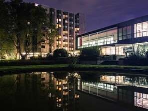 Un edificio moderno con fachada de vidrio, reflejado en el agua y un árbol en primer plano.