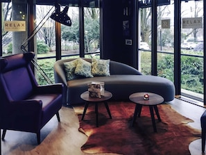 A modern interior with a sofa, armchair, and two small tables, large windows with a view of greenery, and a sign reading 'RELAX'.