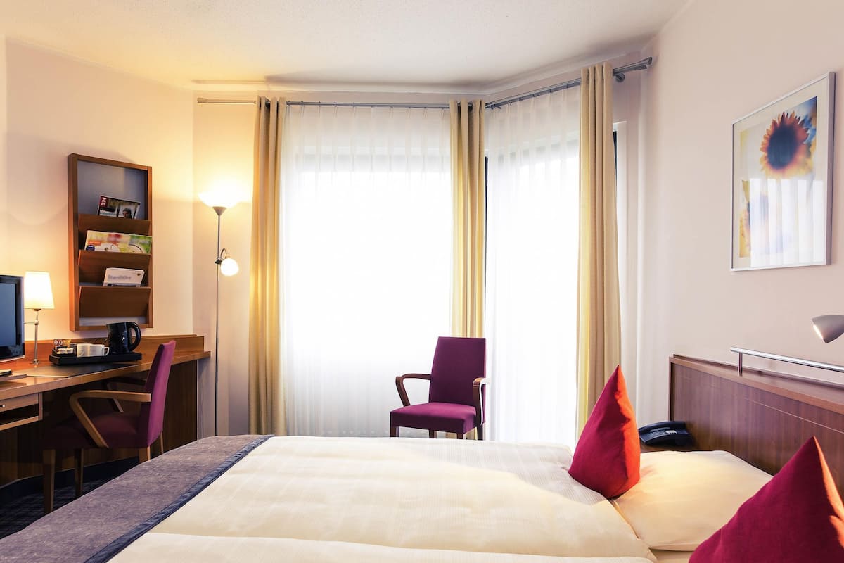 A hotel room with a bed, a desk with a computer, a chair, a bookshelf, and a window with curtains.