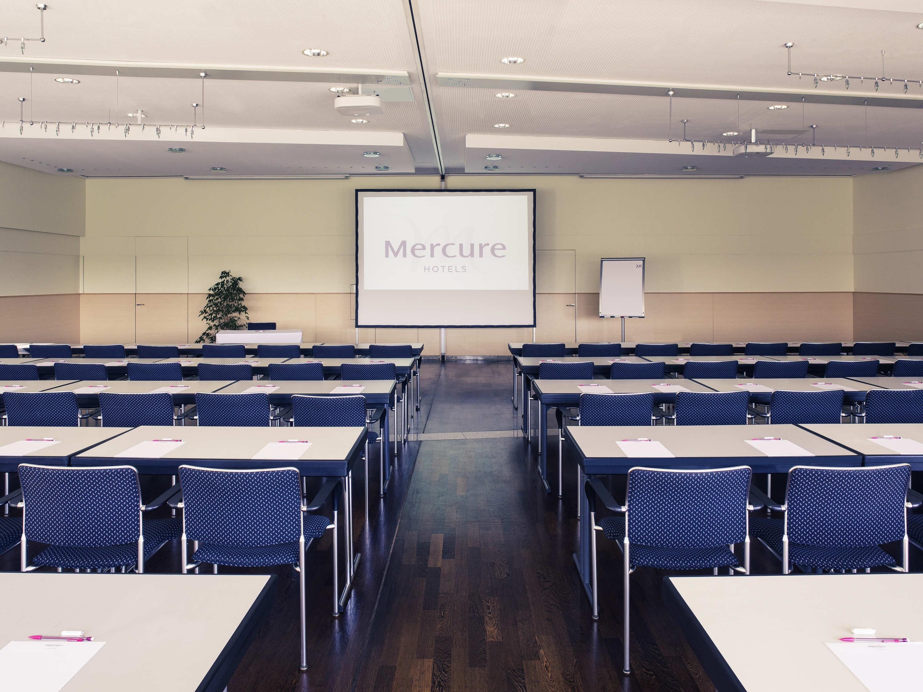 Un salón de conferencias con mesas y sillas dispuestas en filas, una pantalla de proyector y una pizarra.