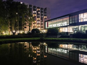 Een modern gebouw met een glazen gevel, een spiegelbeeld in het water, en een verzorgde tuin.