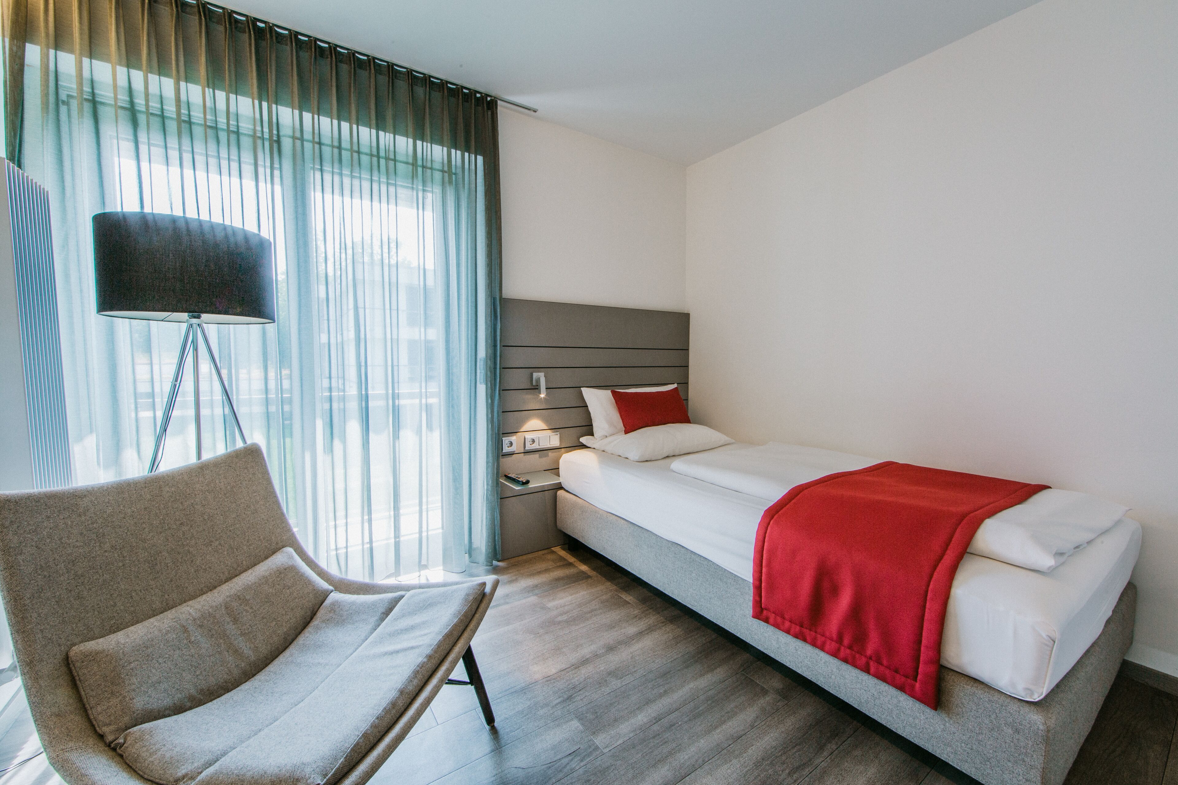 A modern hotel room with a single bed, a bedside table, a lamp, and a chair.