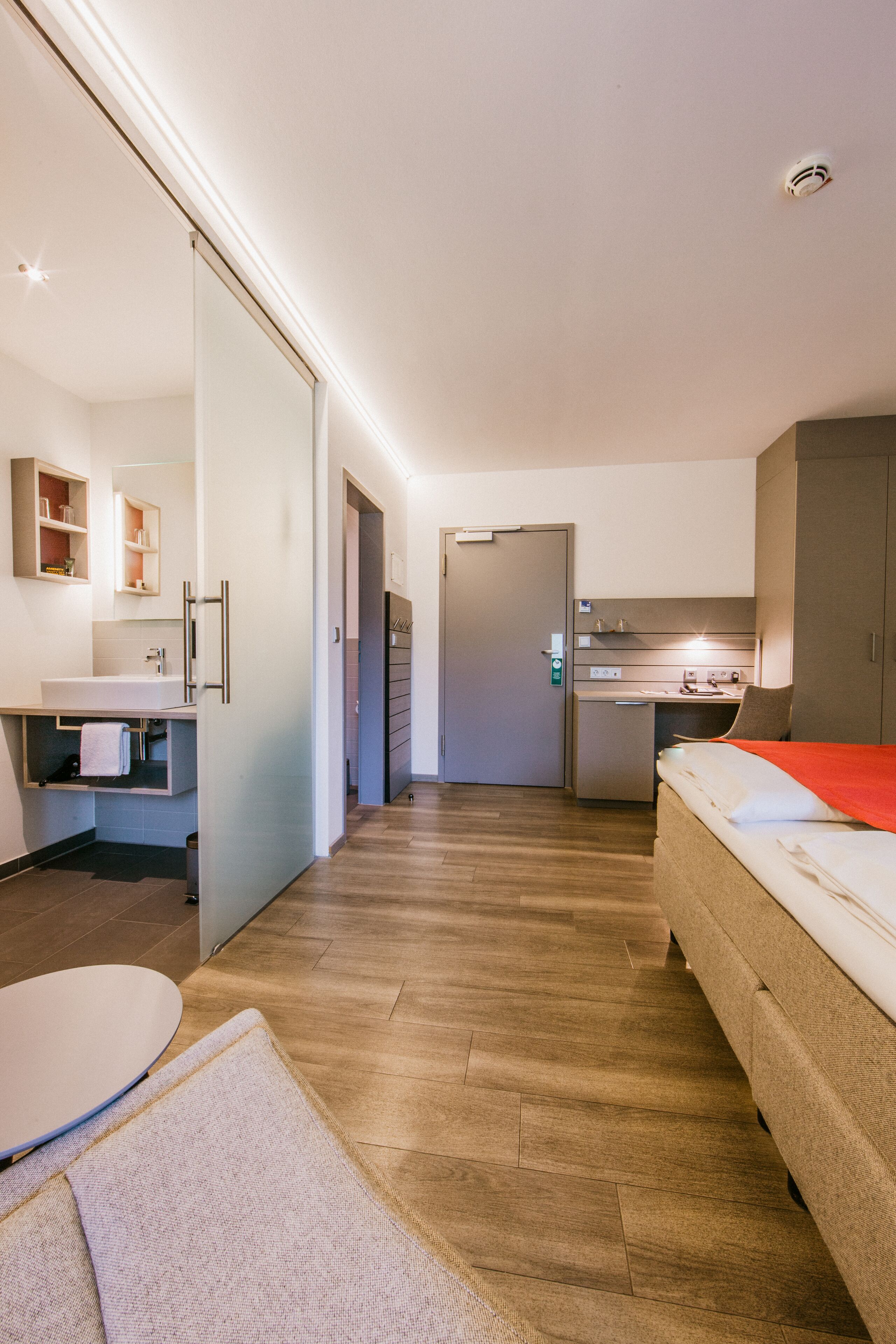 A modern hotel room with a bed, a desk, a kitchenette, and a bathroom.