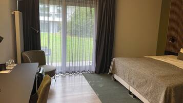 Habitación de hotel con cama, escritorio y silla, una mesa con vasos, y vista a un jardín verde por la ventana.