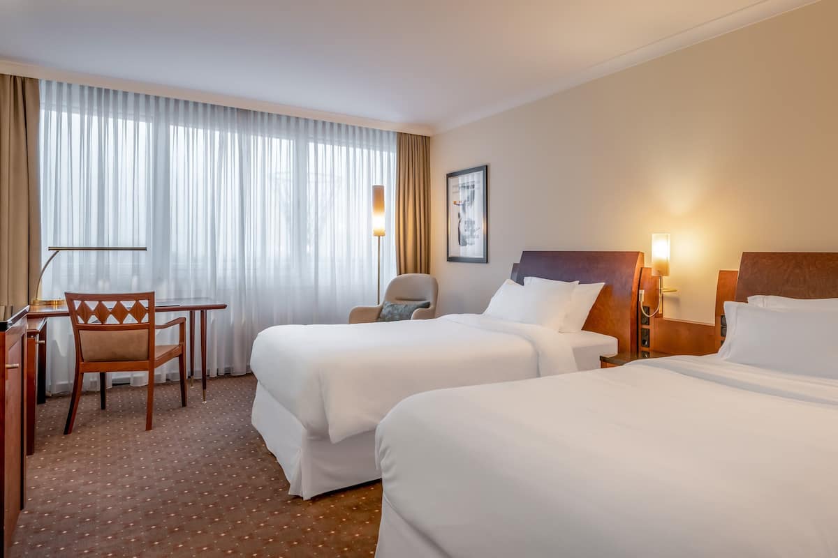 A hotel room with two beds, a desk, a chair, and a large window with curtains.