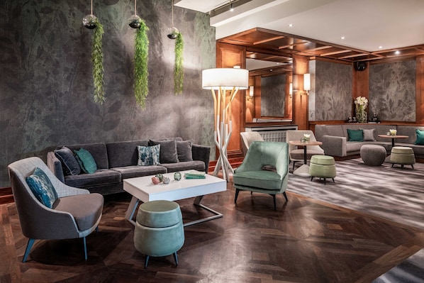 A modern hotel lobby with a dark wood floor, a grey sofa, a white coffee table, and green plants on the wall.