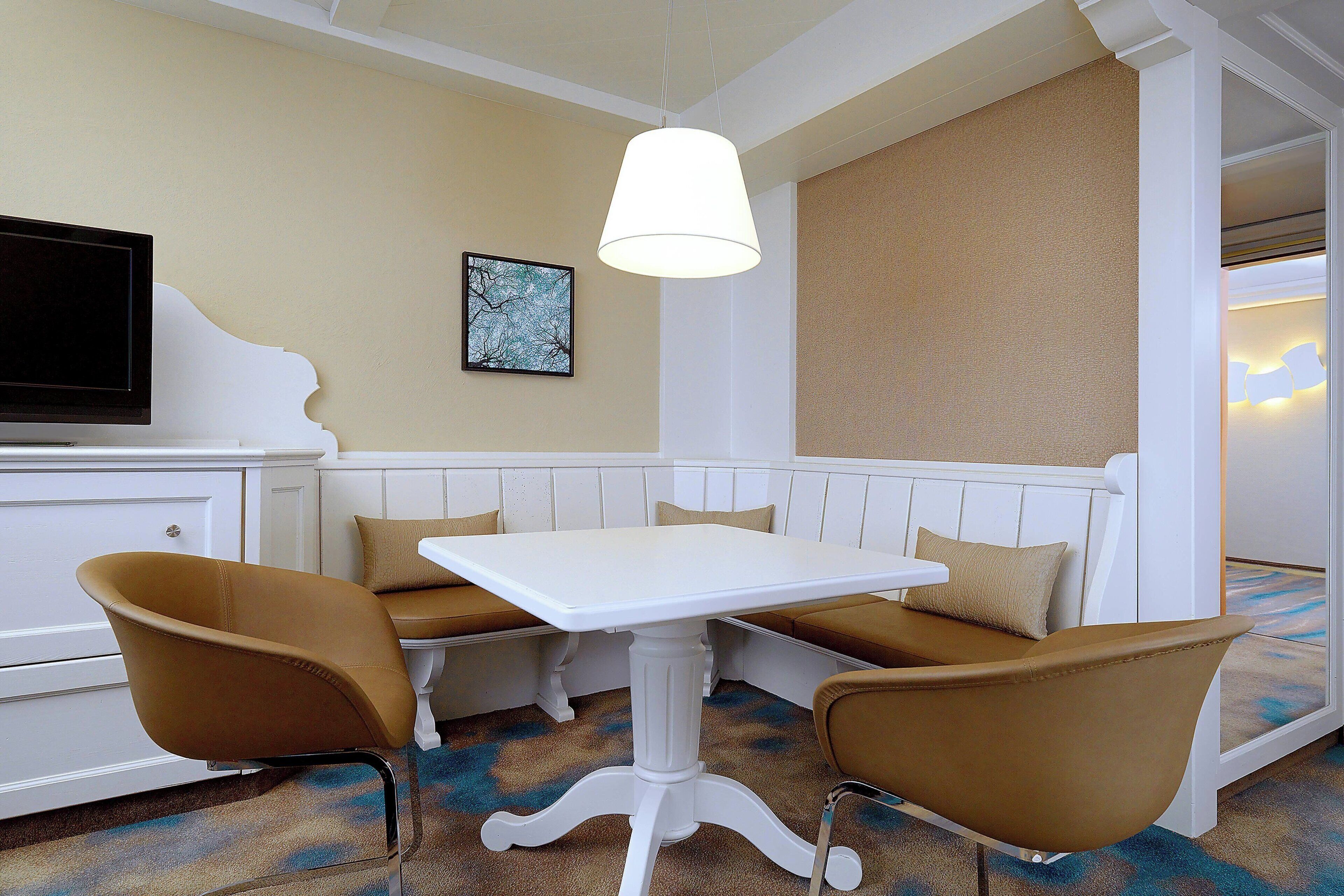 Una habitación con una mesa blanca, dos sillas marrones, un banco blanco, un televisor y un cuadro en la pared.