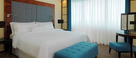 A hotel room with a large bed, a blue bench, a desk with a chair, and a large window with curtains.