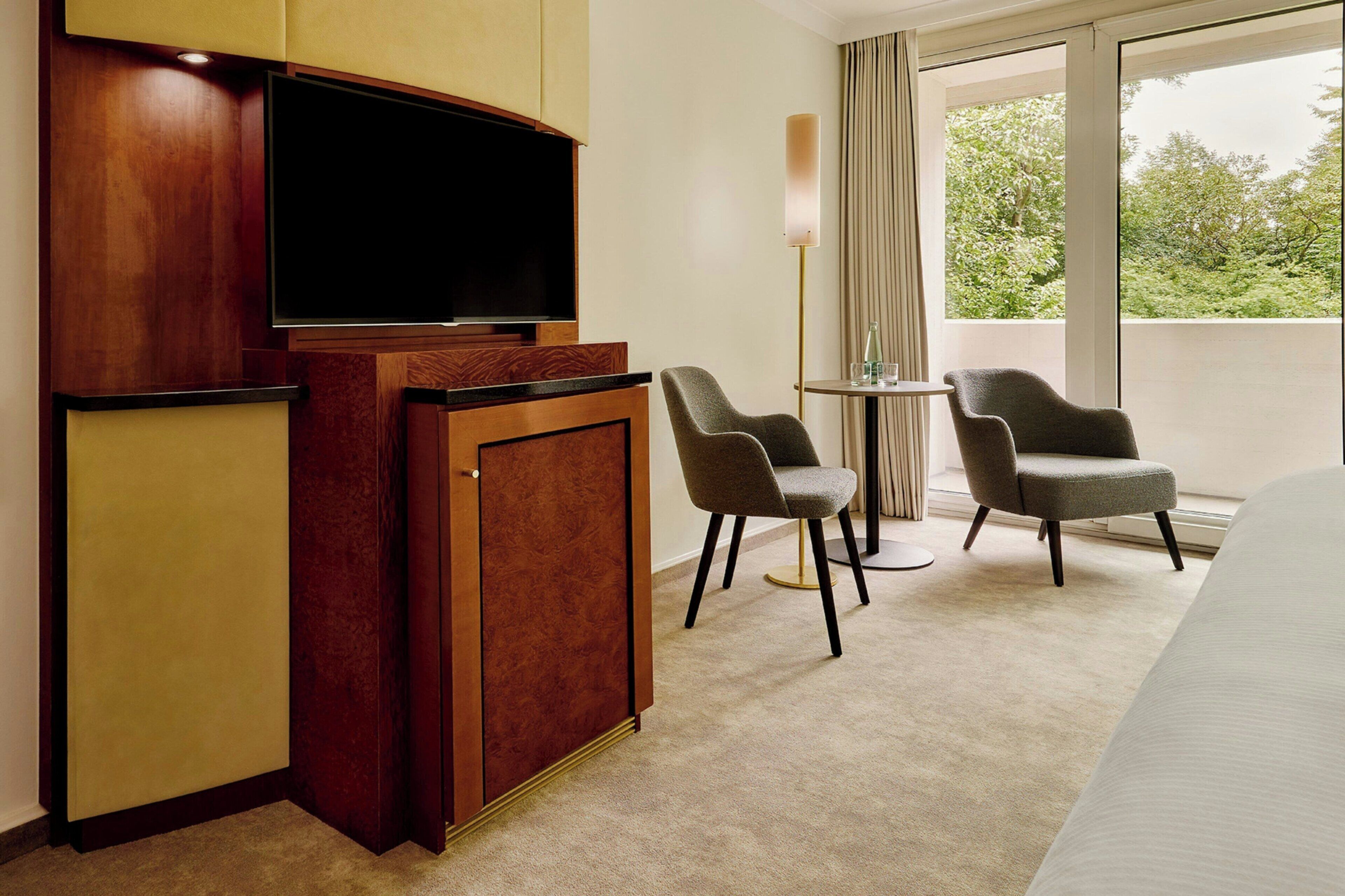 A hotel room with a flat-screen TV, a wooden desk, two chairs, a small round table, and a large window with a view of greenery.