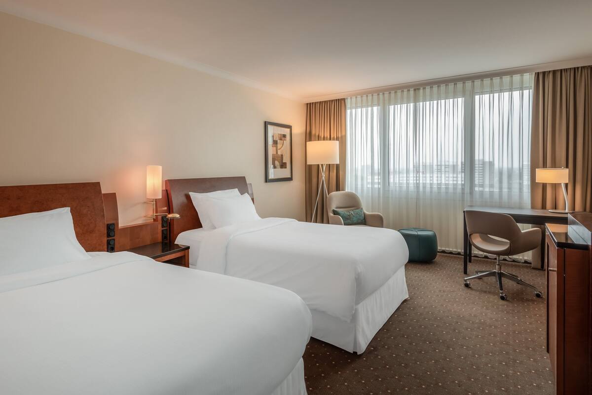 A hotel room with two beds, a desk, a chair, and a large window with curtains.