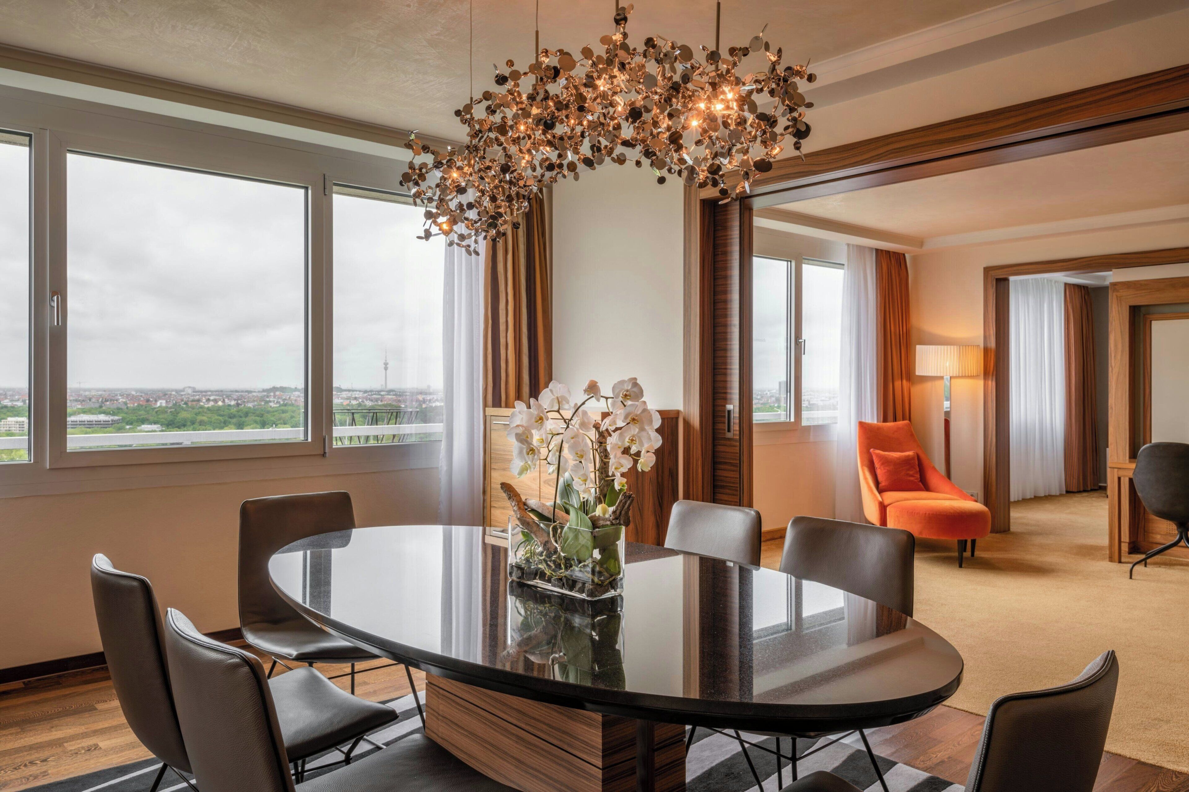 A modern hotel room with a round dining table, chairs, a chandelier, and a view of the cityscape.
