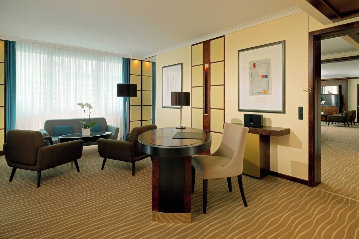 A hotel room with a round dining table, two armchairs, a sofa, a TV, and artwork on the walls.