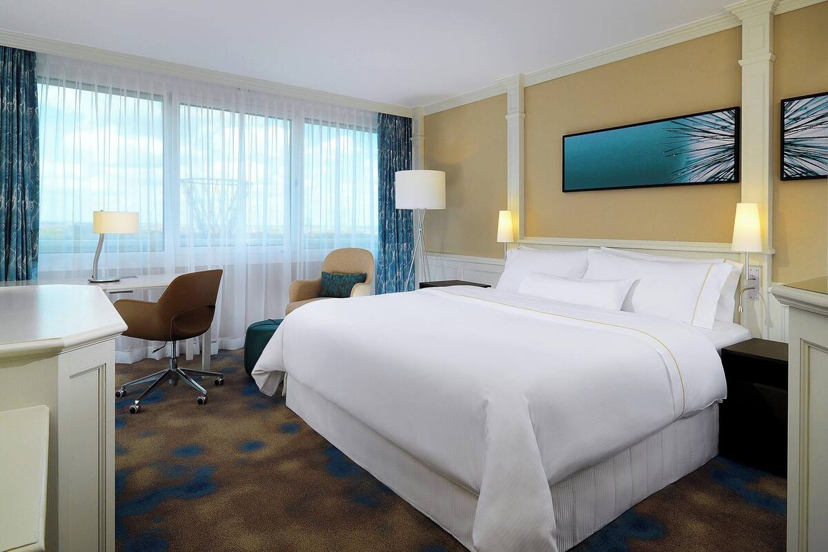 A hotel room with a large bed, a desk, a chair, and a view of the outdoors through sheer curtains.